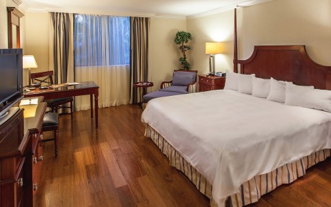 bedroom with wood floors, white bed, lounge chair, desk and chair and dresser with tv 
