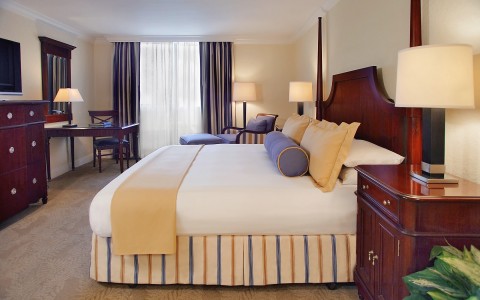 bedroom with white bed, yellow and blue accents, dresser with tv, desk with chair, lounge chair in corner 